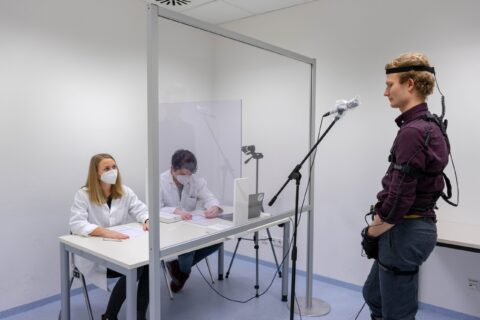 Eine Person mit Messgeräten am Körper steht an einem Mikrofon. Hinter dem Mikrofon sitzen an einem Tisch zwei Personen mit Mundschutz und weißen Mänteln.
