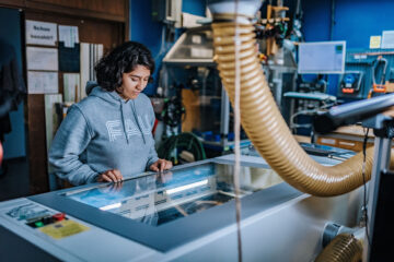 Eine junge Frau trägt einen grauen Pullover und blickt auf ein technisches Gerät, das vor ihr steht.