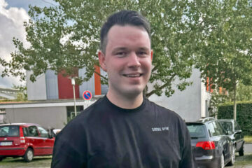 Eine Person mit kurzen, dunklen Haaren steht vor einem Baum. Die Person trägt ein dunkles T-Shirt.