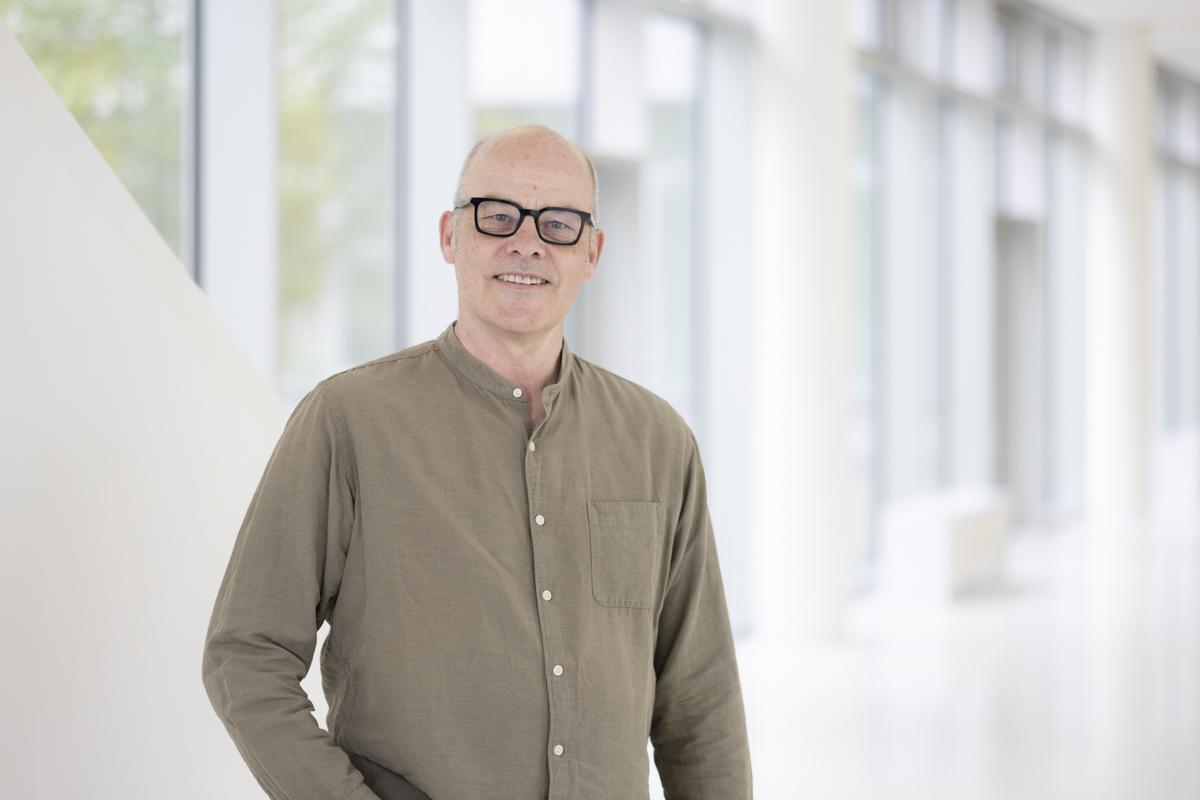 Eine Person mit einem hellbraunen Hemd steht vor einer weißen Wand mit Fenstern. Die Person hat eine Glatze, trägt eine schwarze Brille und schaut lächelnd in die Kamera.