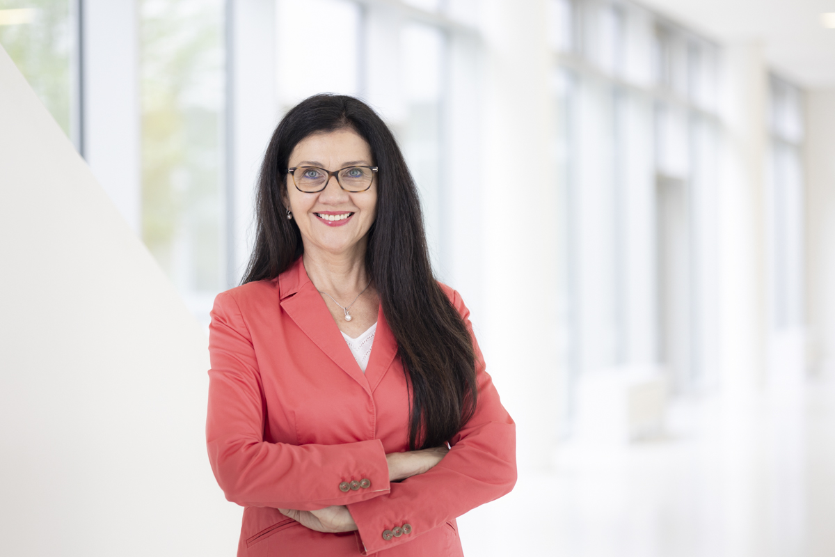 Eine Person mit roter Jacke über einem weißen Shirt steht mit verschränkten Armen vor einer weißen Wand mit Fenstern. Die Person hat lange schwarze Haare, trägt eine Brille und blickt lächelnd in die Kamera.