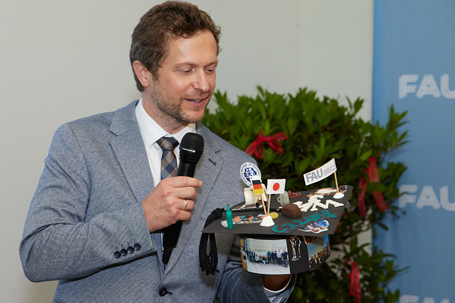 Eine Person im hellen Anzug hält in einer Hand einen Doktorhut und in der andern Hand ein Mikrofon.