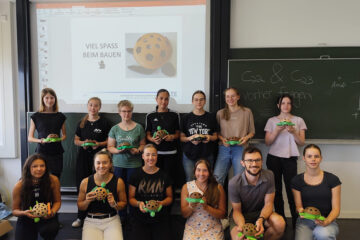 In zwei Reihen stehen bzw. knien mehrere Personen vor einer Wand. Die Personen halten eine selbstgebastelte Schildkröte in der Hand. Auf die Wand ist ein Bild projiziert, auf dem steht "Viel Spaß am bauen".