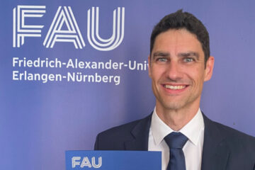 Eine Person mit dunklen, kurzen Haaren steht vor einer blauen Wand auf die in weißen Buchstagen das Logo der FAU gedruckt ist. Die Person trägt ein weißes Hemd, eine blaue Krawatte, eine blaue Anzugjacke und blickt freundlich lachend in die Kamera.
