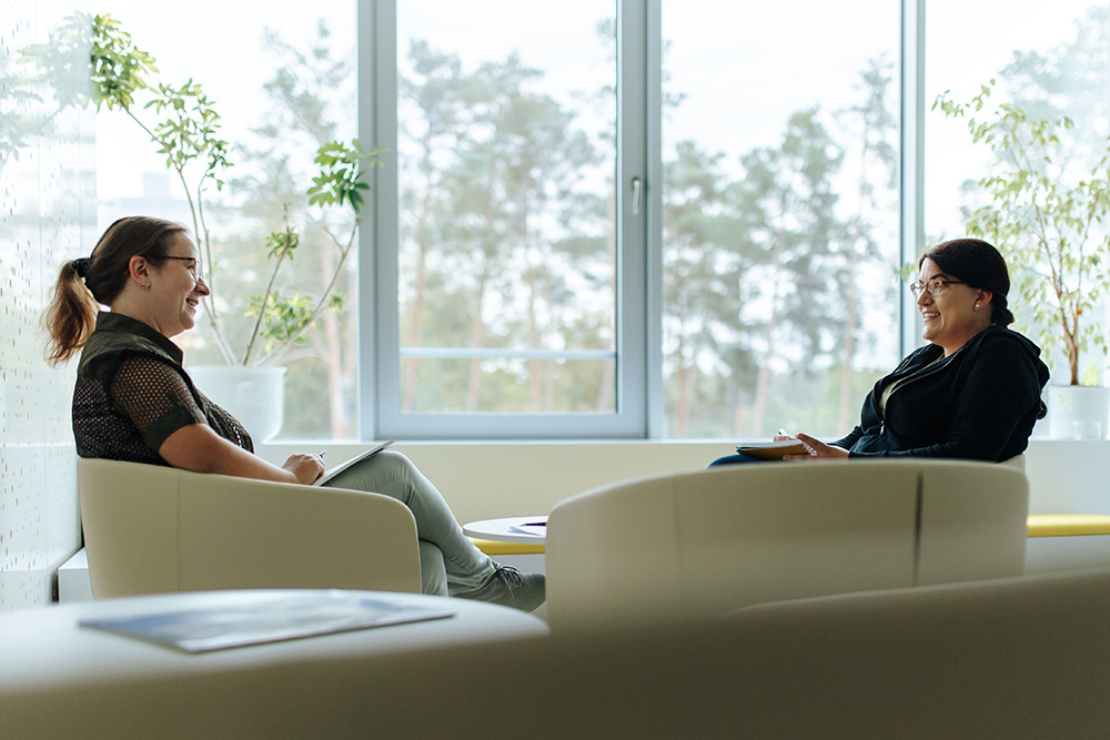 Vor einem großen Fenster sitzen sich 2 Personen in Sesseln gegenüber. Sie halten beide Papier und Stift in der Hand und blicken sich freundlich an. Die Personen haben beide dunkle Haare, tragen ein dunkles Oberteil und eine Brille.