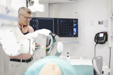 Eine Person betrachtet ein Krankenhausbett, an dem ein Roboterarm und im Hintergrund Bildschirme zu sehen sind.