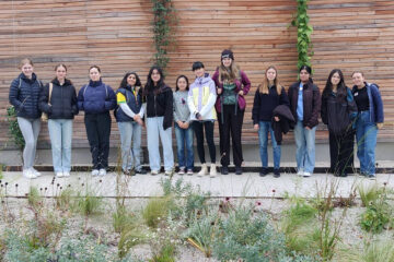 12 junge Frauen stehen nebeneinander vor einer Holzwand und blicken in die Kamera.