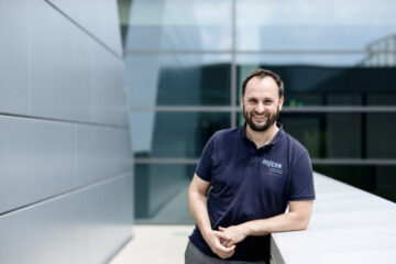 Eine Person steht vor einer Glaswand und lehnt sich mit dem einem Arm an einer Mauer an. Die Person lächelt in die Kamera.