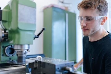 Eine Person mit hellen, kurzen Haaren und einer Brille schaut auf eine Maschine. Die Maschine ist hellgrün und hat metall-farbene und schwarze Teile.