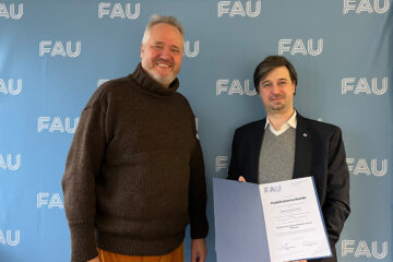 Zwei Personen stehen vor einer hellblauen Wand, auf die mehrmals die weißen Buchstaben FAU gedruckt sind. Die Personen blicken freundlich in die Kamera und halten eine blaue Mappe mit einer weißen Urkunde zwischen sich.