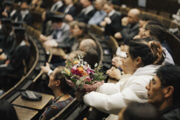 Mehrere Personen sitzen in einem Hörsaal. Der Fokus liegt auf einer Frau mit weißer Jacke und einem bunten Blumenstrauß in der Hand.