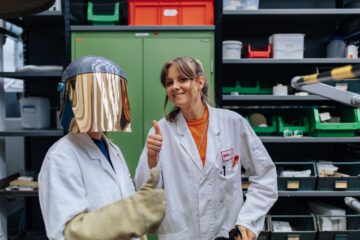 Zwei Personen in weißen Mänteln stehen vor einem grünen Schrank. Die eine Person hat eine glänzende Maske vor dem Gesicht, die andere lächelt in die Kamera und hält ihren Daumen nach oben.