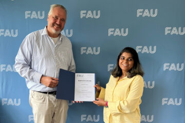 Zwei Personen stehen vor einer hellblauen Wand, auf die mehrmals die weißen Buchstaben FAU gedruckt sind. Die Personen blicken freundlich in die Kamera und halten eine blaue Mappe mit einer weißen Urkunde zwischen sich.