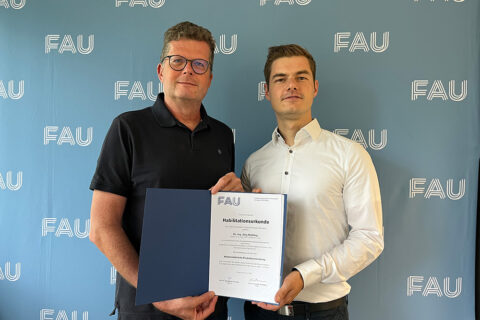 Zwei Personen stehen vor einer hellblauen Wand, auf die mehrmals die weißen Buchstaben FAU gedruckt sind. Die Personen blicken freundlich in die Kamera und halten eine blaue Mappe mit einer weißen Urkunde zwischen sich.