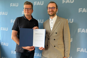 Zwei Personen, eine Person mit dunklem, kurzärmlichen Hemd und eine Person mit hellkarierter Jacke, stehen vor einer hellblauen Wand, auf die mehrmals die weißen Buchstaben FAU gedruckt sind. Die Personen blicken freundlich in die Kamera und halten eine blaue Mappe mit einer weißen Urkunde zwischen sich.