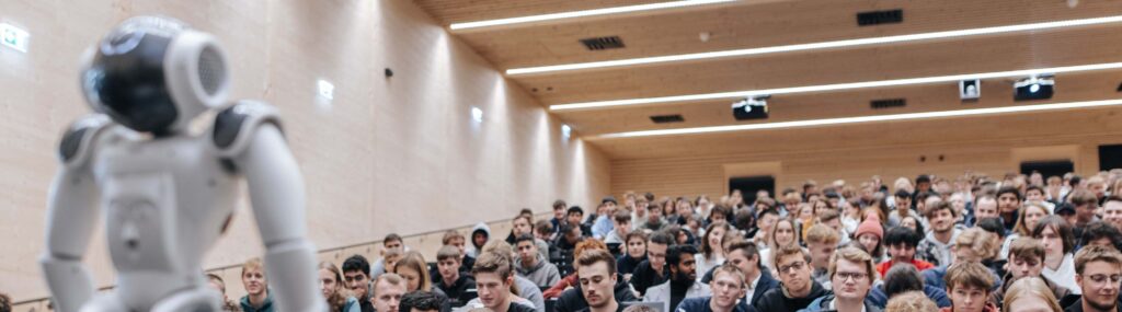 Ein Roboter steht vorne im Hörsaal einer Universität und blickt auf Sitzreihen voller Studierender.
