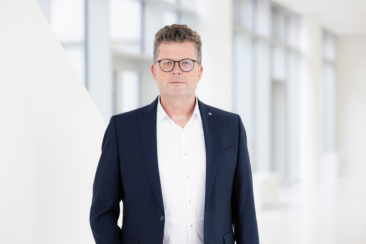 Eine Person mit kurzen, lockigen Haaren und Brille schaut in die Kamera. Die Person trägt ein weißes Hemd und eine blaue Jacke darüber.