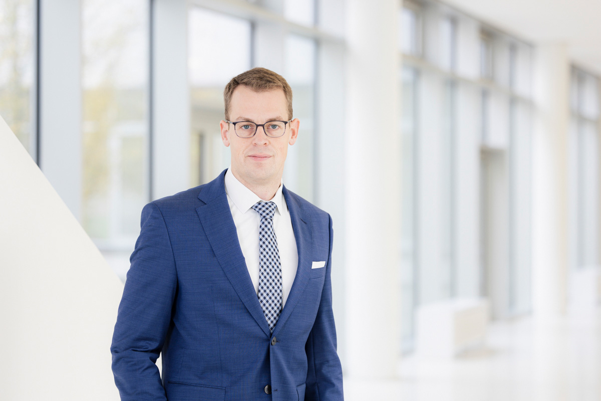 Eine Person mit kurzen, braunen Haaren und Brille schaut in die Kamera. Die Person trägt ein weißes Hemd mit einer blau-weiß karierten Krawatte und eine blaue Jacke darüber.