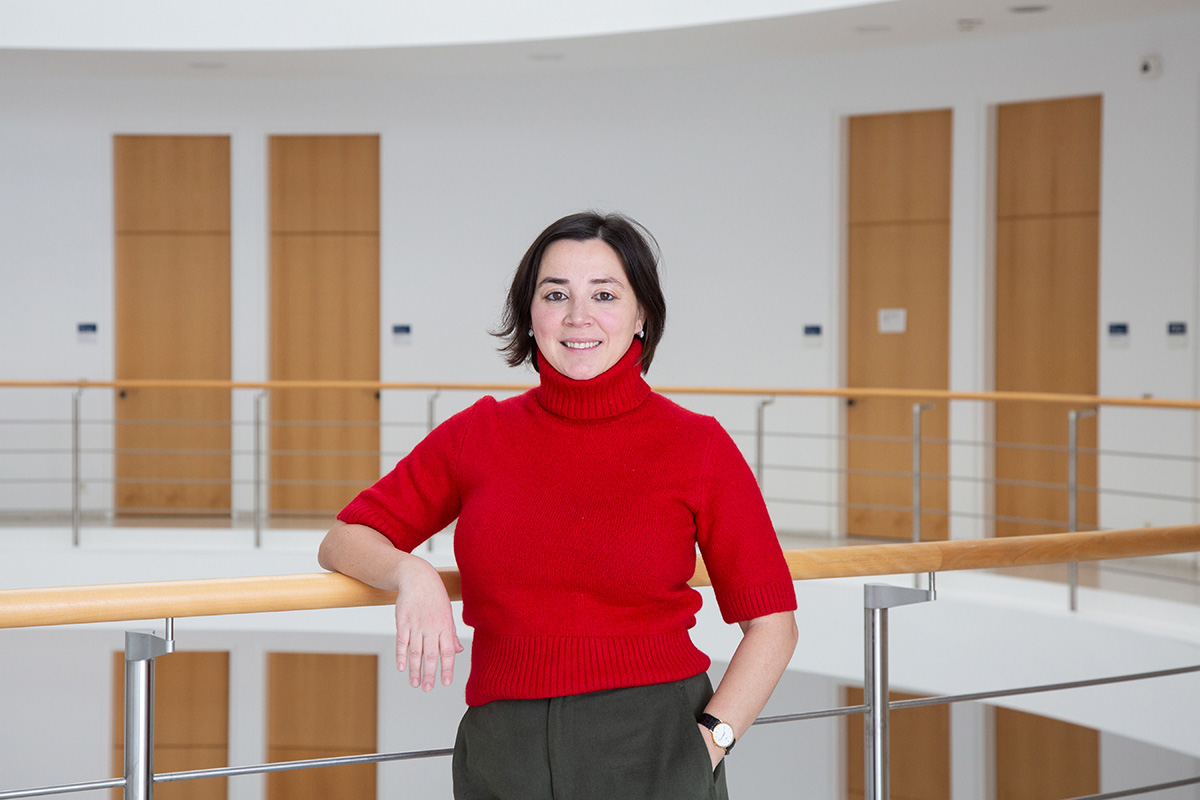 Eine Person mit mittellangen dunklen Haaren schaut lächelnd in die Kamera. Die Person trägt einen kurzärmligen roten Pullover.