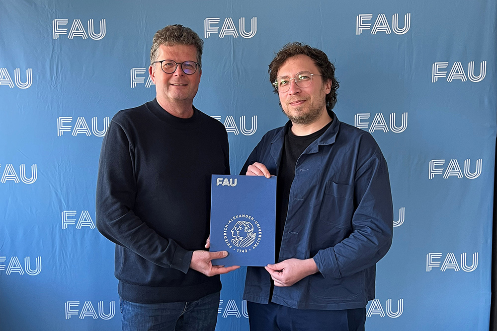 Zwei Personen stehen nebeneinander und blicken freundlich in die Kamera. Sie halten vor sich eine blaue Mappe.